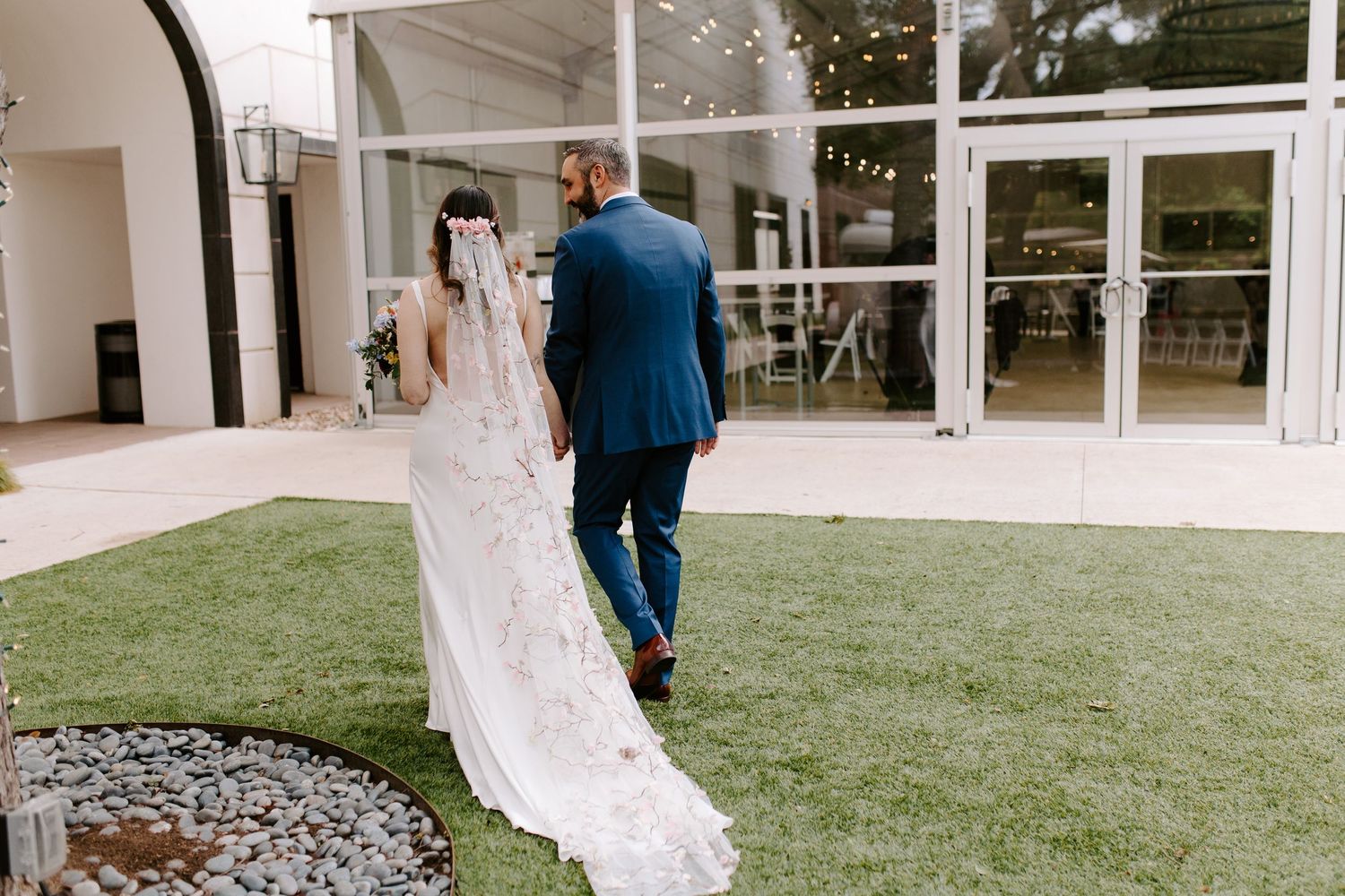 A couple in wedding attire walking on lush green grass by a modern glass building with a flowing lace train.