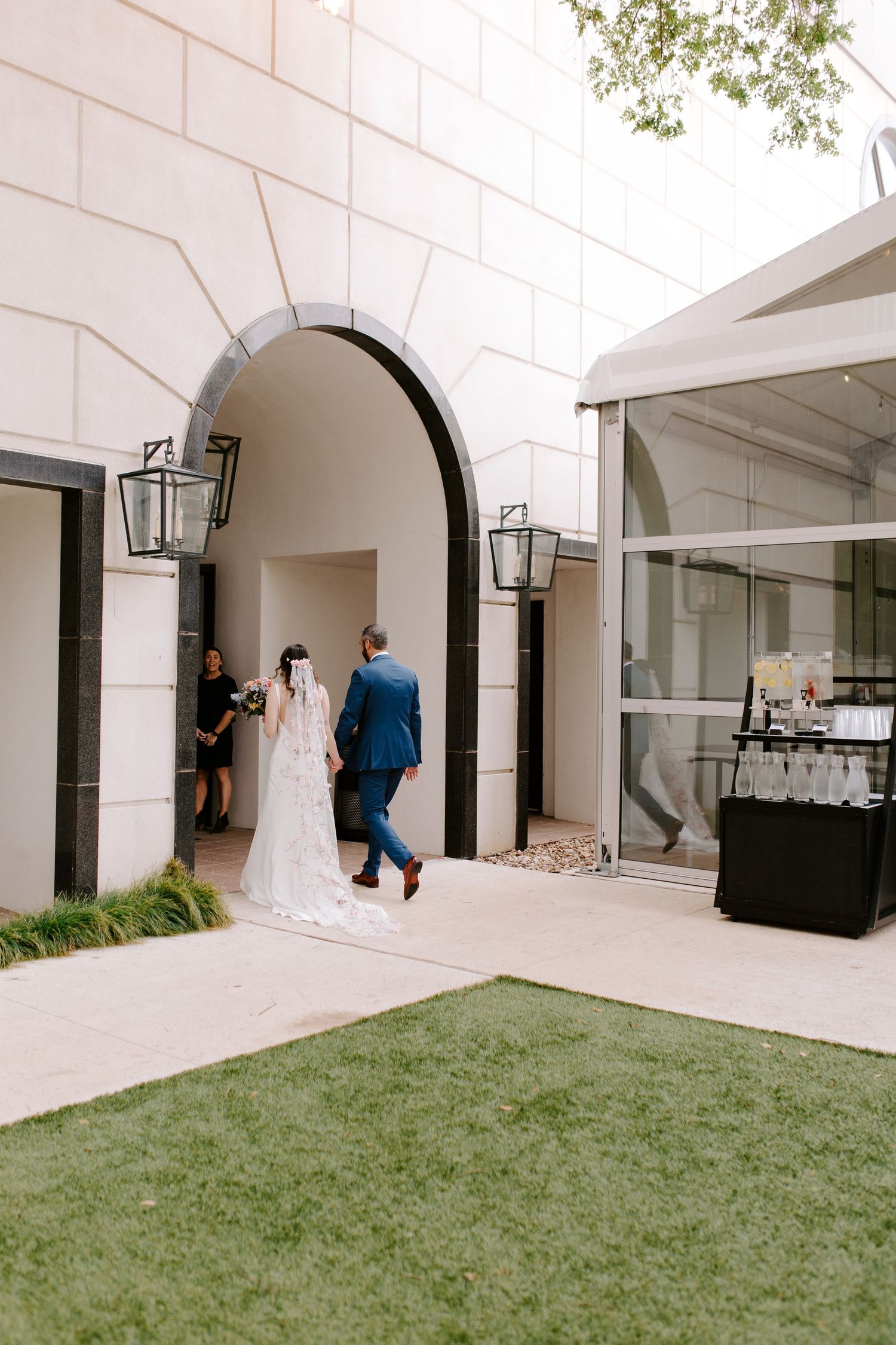 Wedding party walking through stone archway with white exterior walls and black lanterns.