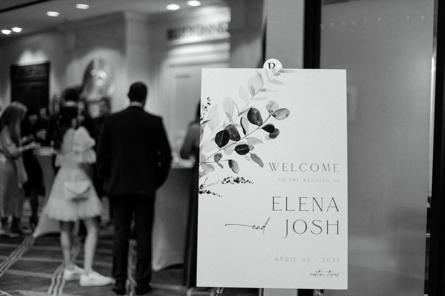 Black and white welcome sign for Elena and Josh's wedding with floral design displayed in event space.