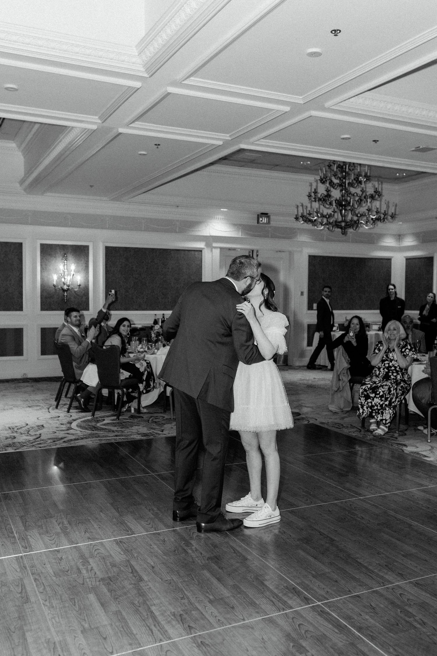 Black and white photo of a couple dancing in a ballroom with hardwood floors and elegant chandeliers.