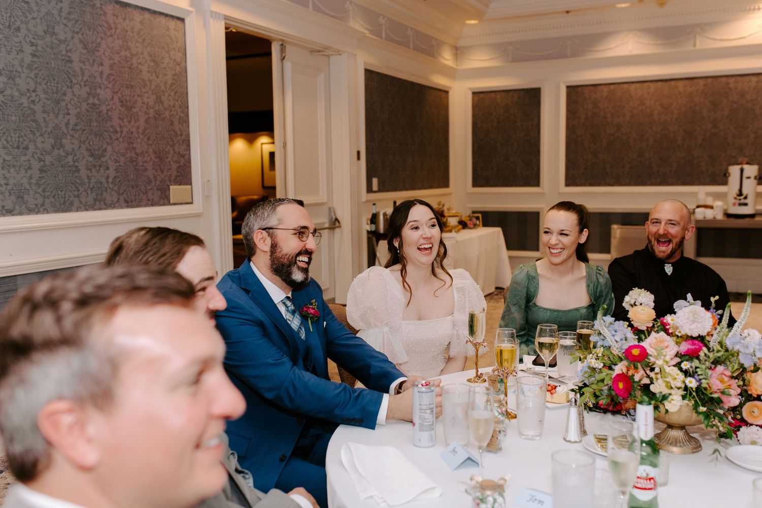 Wedding reception guests laugh together over dinner at an elegantly decorated table with floral centerpieces.