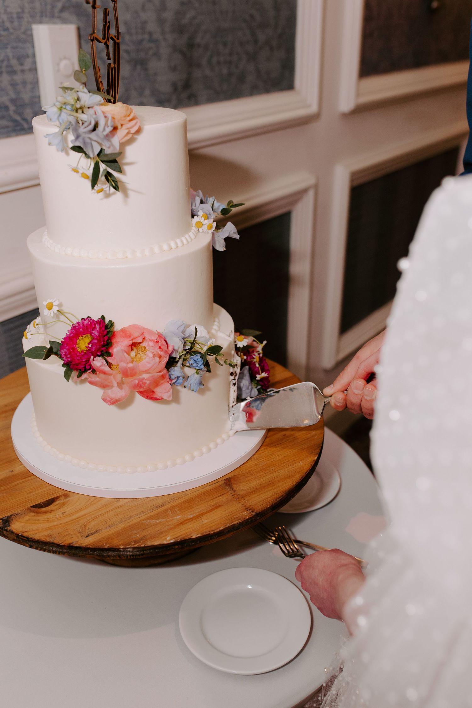 Three-tier white wedding cake decorated with colorful fresh flowers on a wooden cake stand.