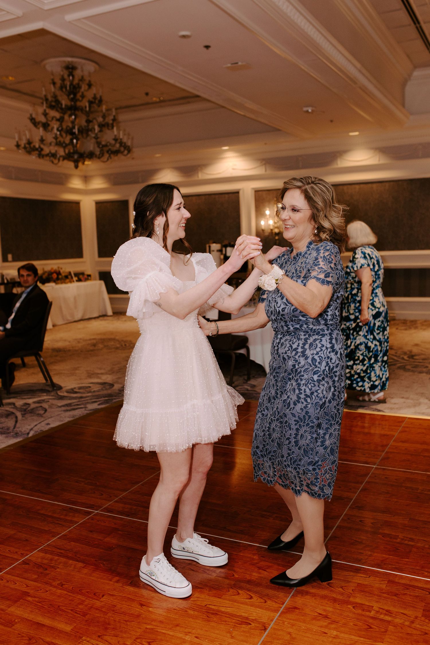 Two guests dancing together on a wooden dance floor at an indoor reception with elegant chandeliers overhead.