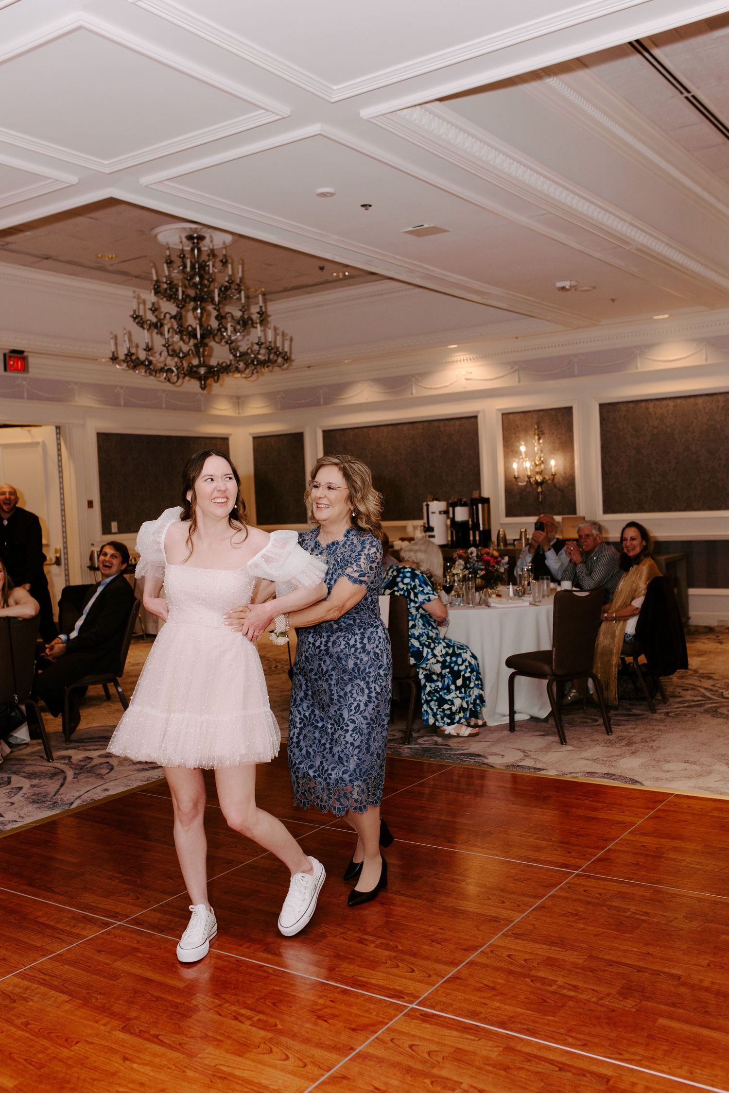 Dancing guests enjoy themselves on a polished hardwood floor under elegant chandeliers at a wedding reception.