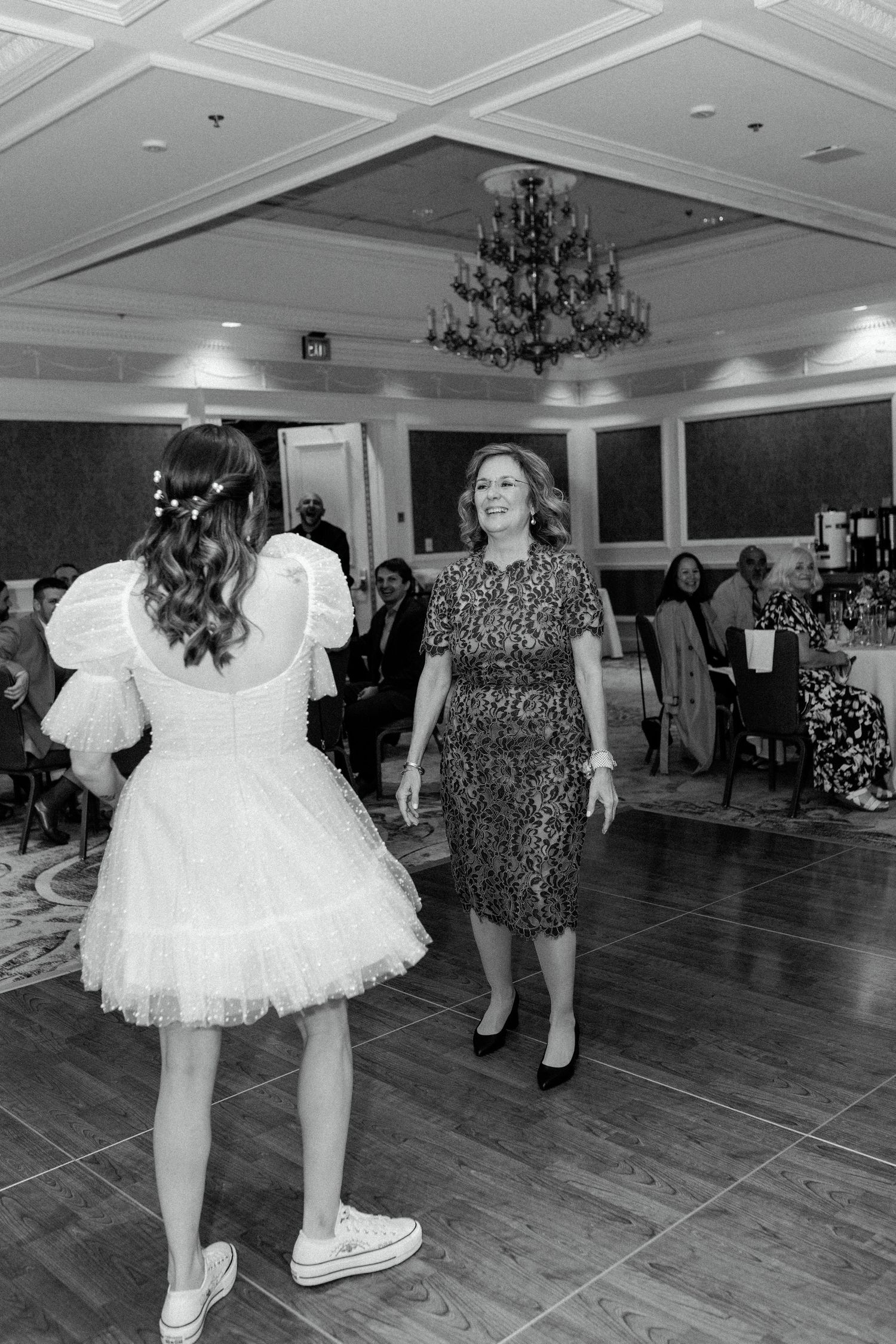 Wedding guests dance on a wooden floor beneath an elegant crystal chandelier.