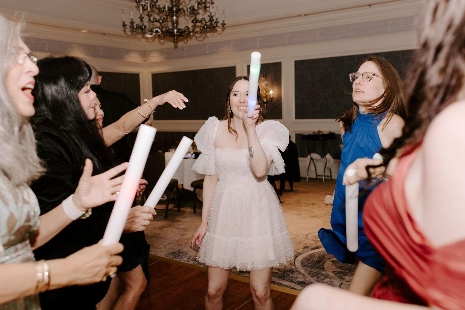 People dancing and celebrating at an elegant indoor reception venue with a chandelier overhead.