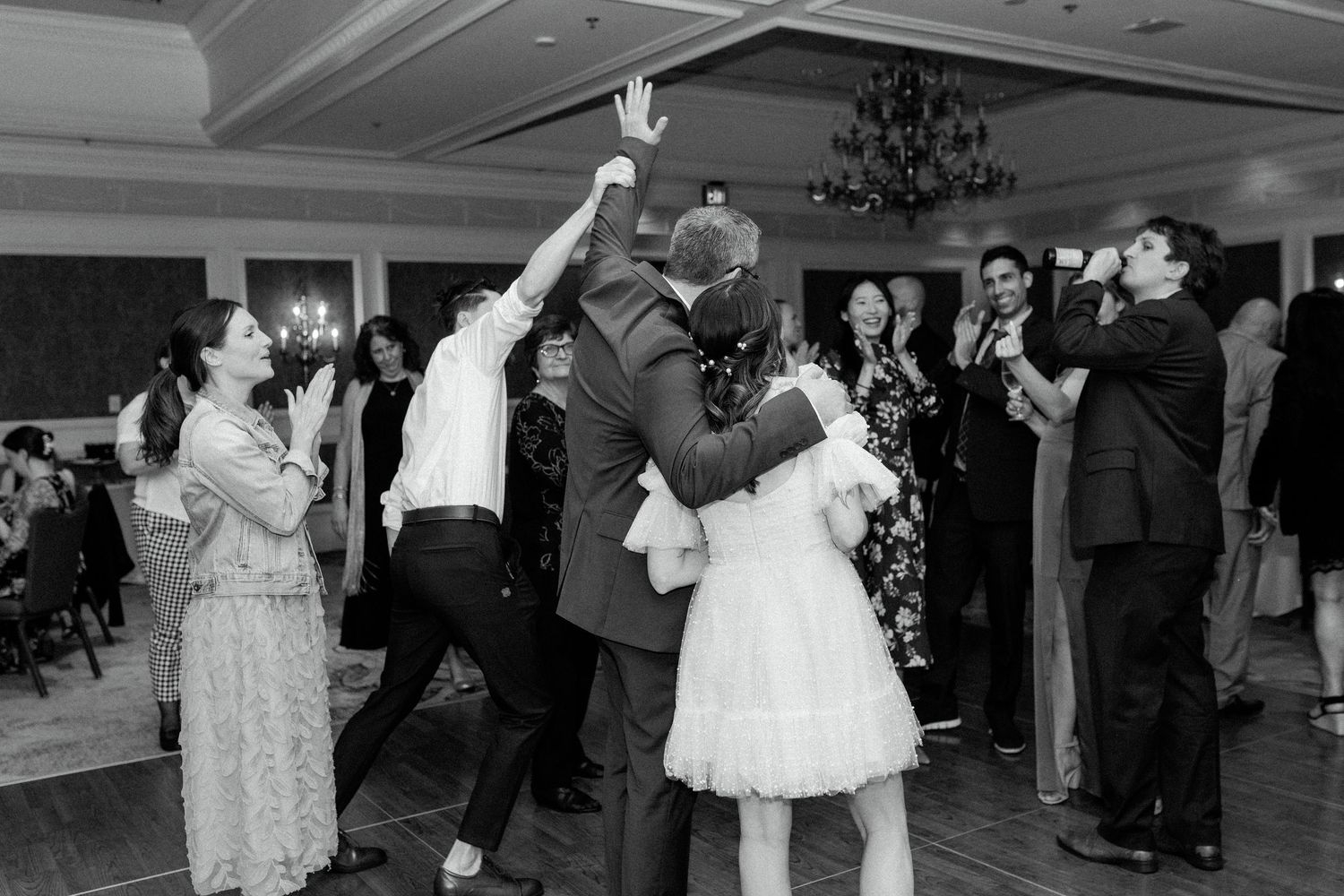 People dancing and celebrating at a wedding reception in a ballroom with a chandelier, captured in black and white.