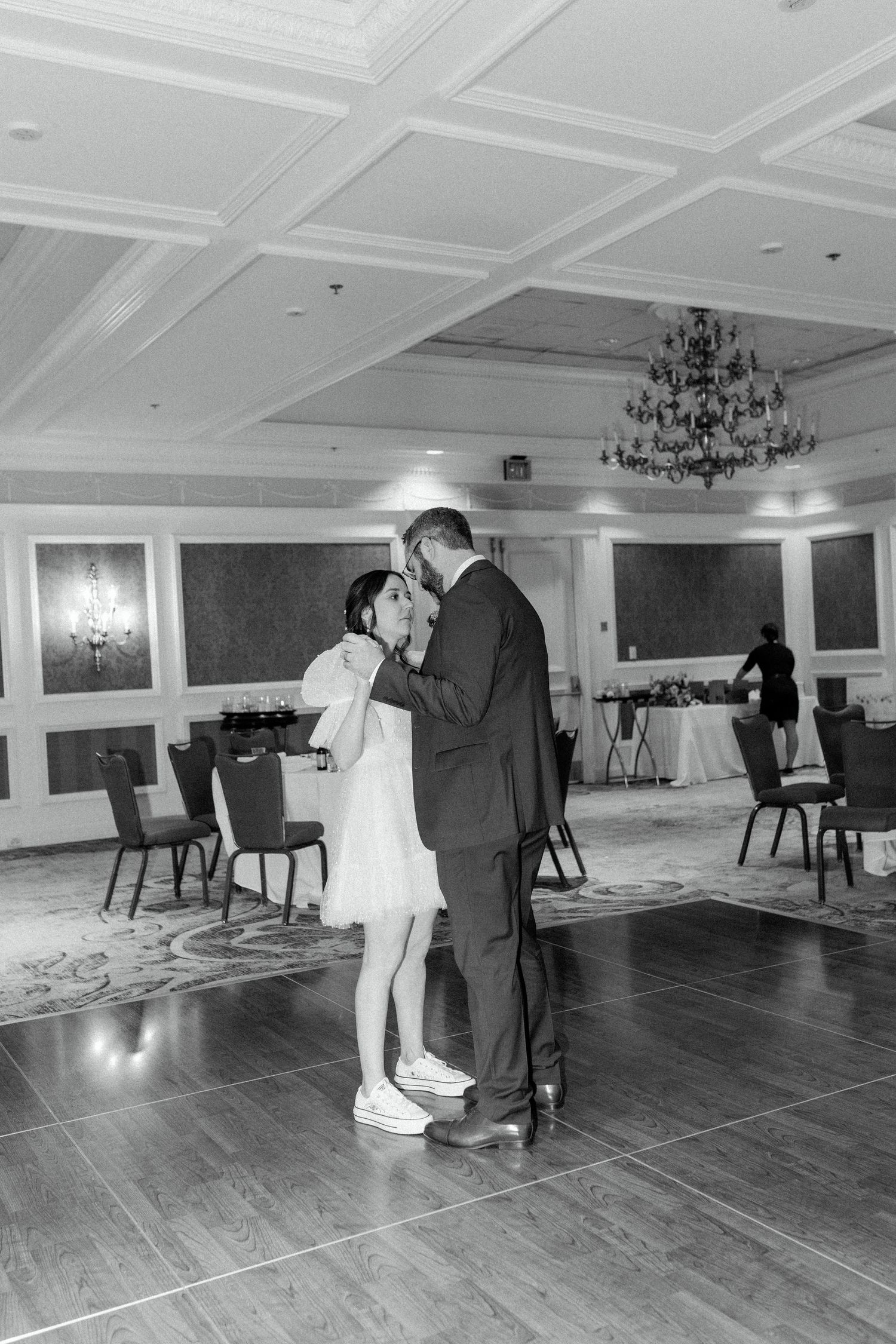 A couple shares a dance together on a hardwood floor in an elegant ballroom with chandeliers and paneled walls.