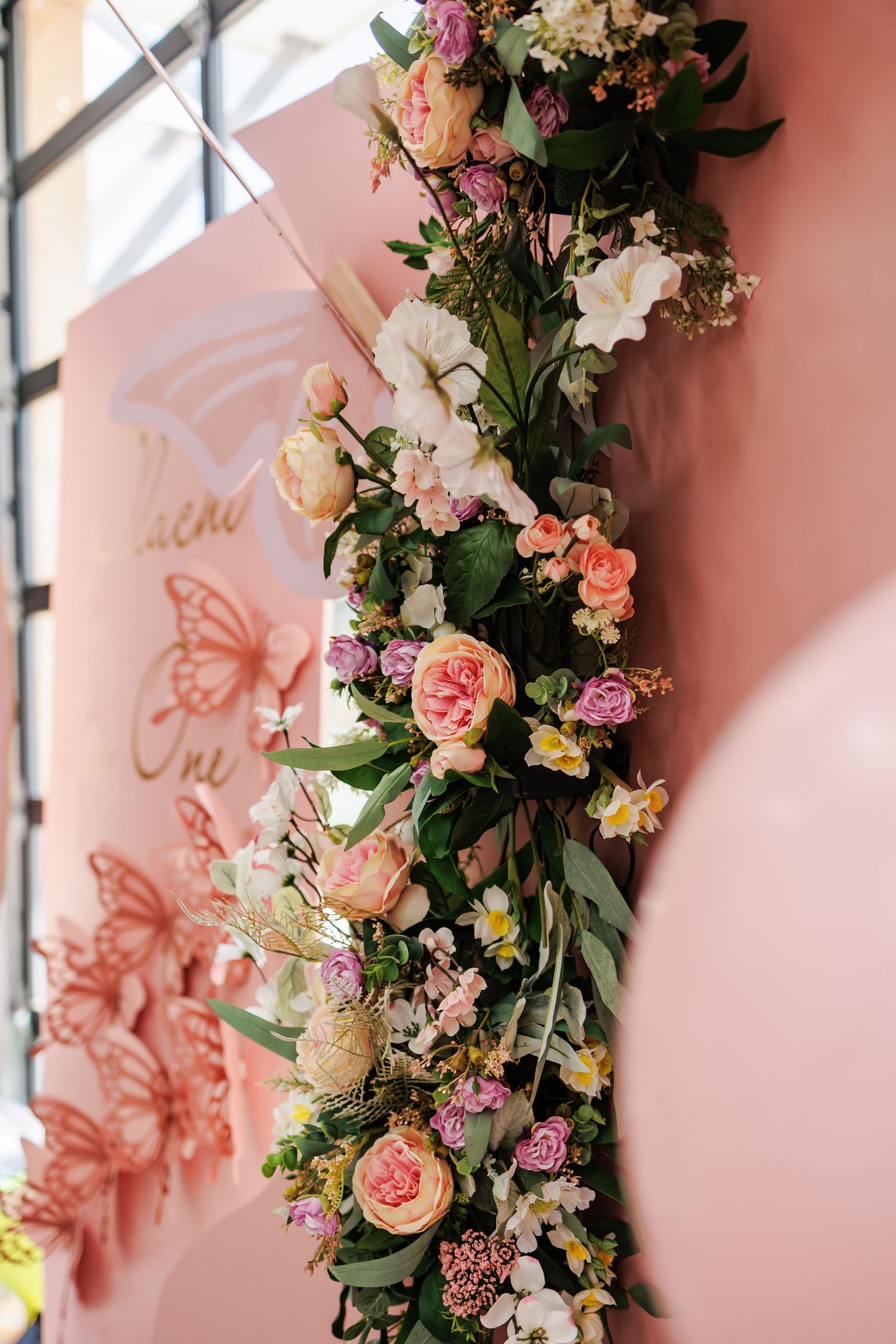 Beautiful floral arrangement with pink roses and mixed flowers against a pink photo backdrop with decorative butterfly decals.