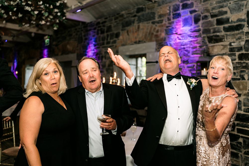 Wedding party guests laughing and celebrating in formal attire at an indoor stone-walled venue with purple lighting.