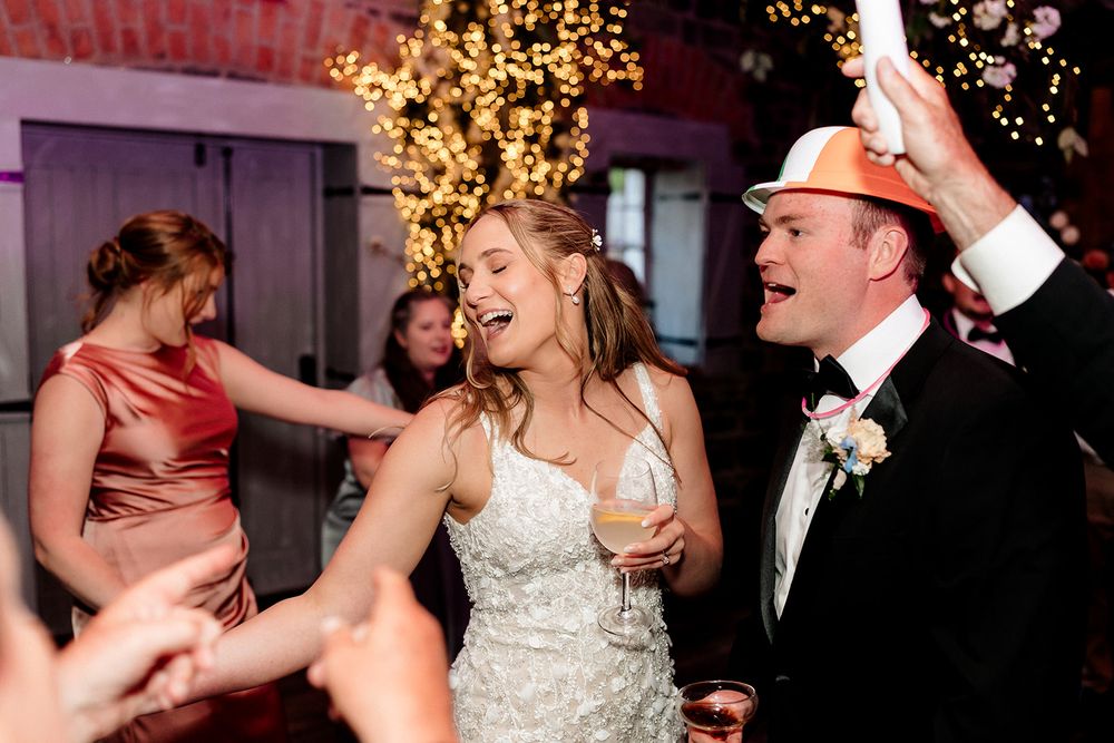 Wedding guests dance joyfully on the dance floor with festive lights and decorations in the background at an evening reception.
