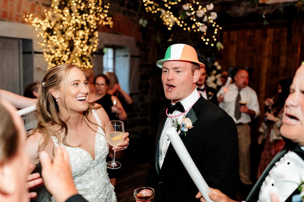 A joyful wedding celebration with people enjoying drinks and dancing under glowing string lights at a rustic venue.
