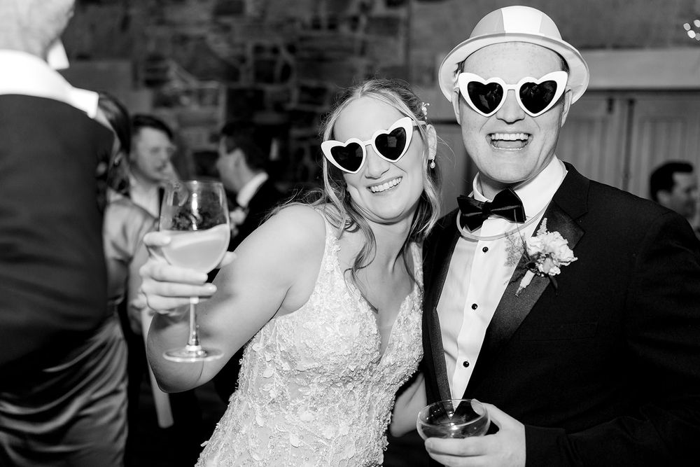 A bride and groom wearing matching heart-shaped sunglasses smile during their wedding reception in black and white.