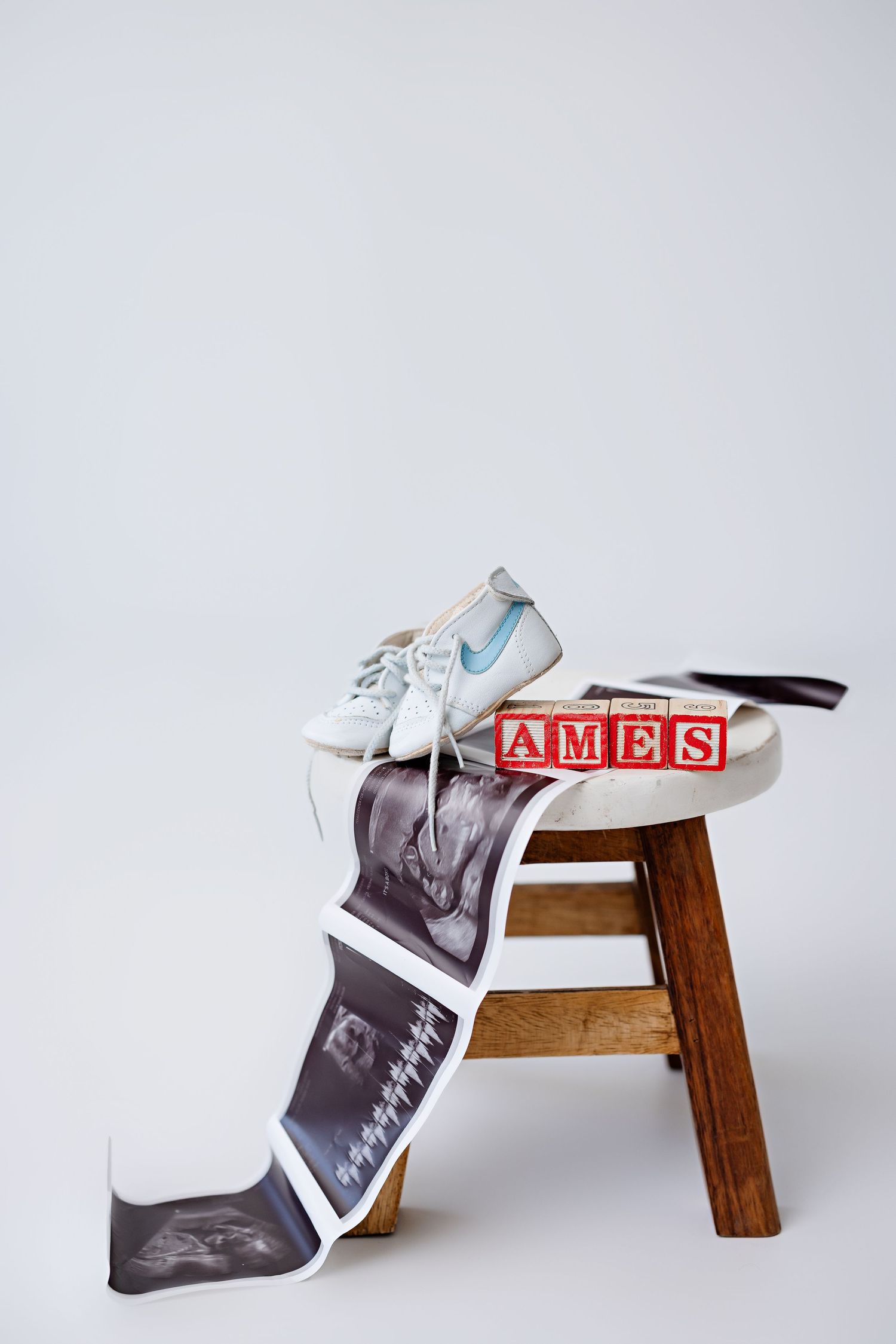 Vintage wooden stool with white baby shoes, alphabet blocks spelling 'AMES', and ultrasound photos draped over it.