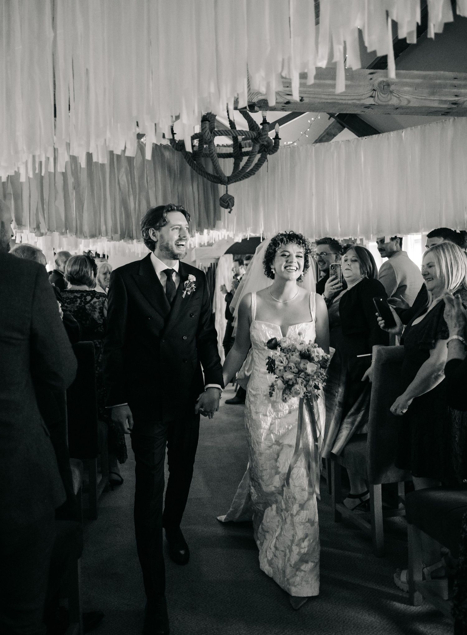 A wedding couple exits through a decorated venue corridor with hanging fabric while guests line the sides in celebration.