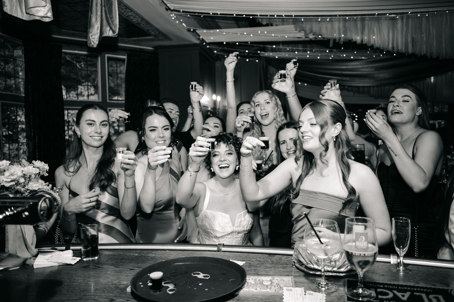 A lively black and white scene of people raising drinks and celebrating at a bar in a joyful party atmosphere.
