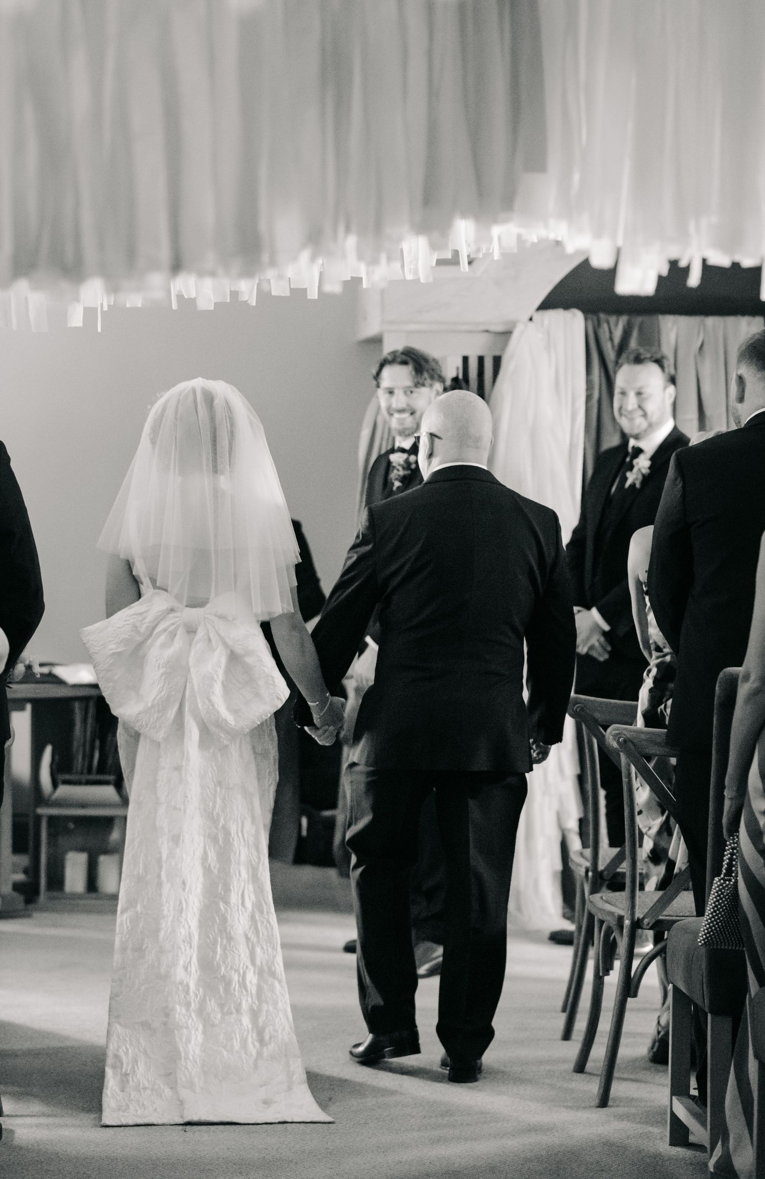 Black and white wedding ceremony photo showing a bride in a lace gown and veil walking down the aisle at a church.