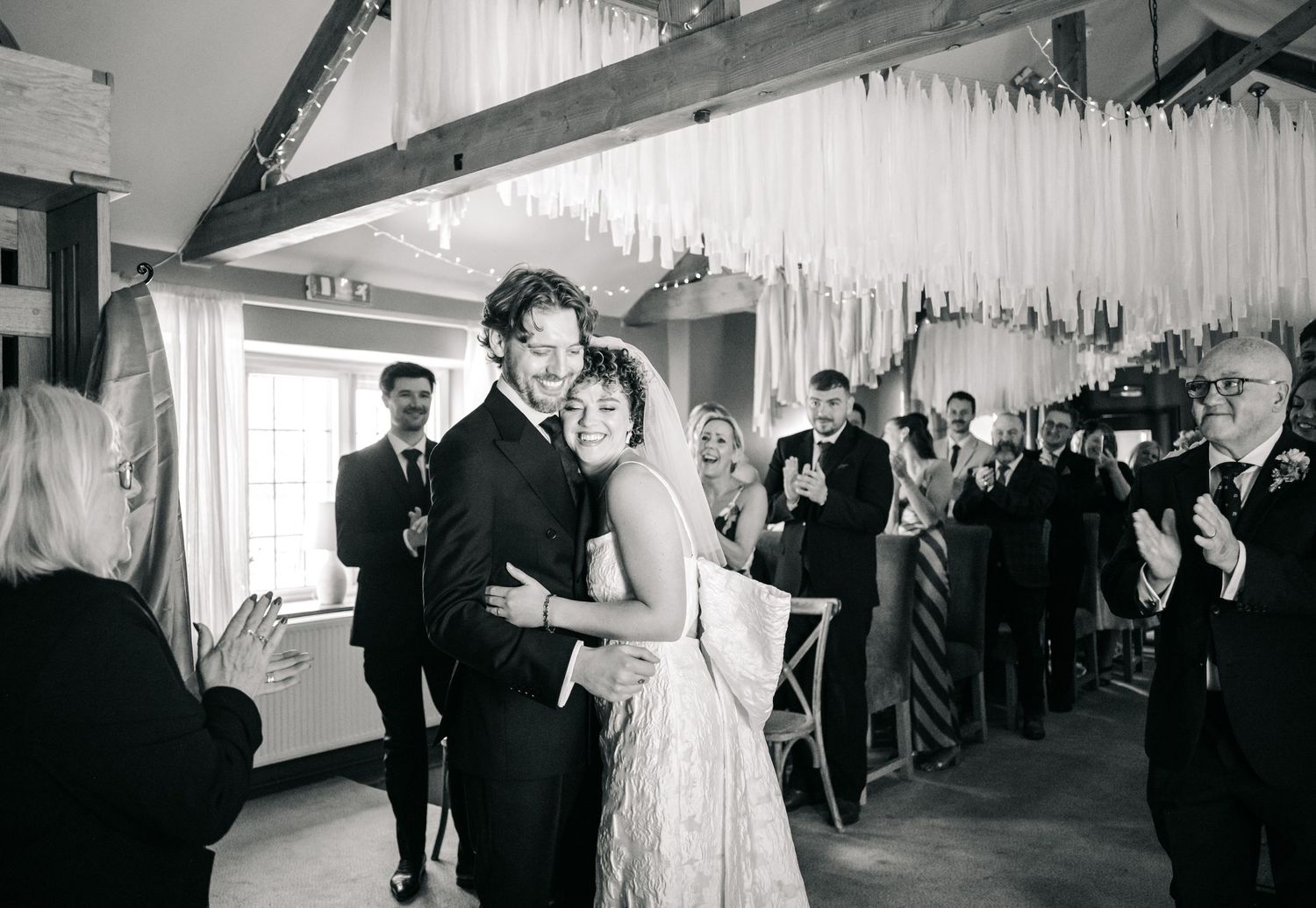 A couple shares their first dance at a wedding reception under decorative hanging white streamers while guests look on.