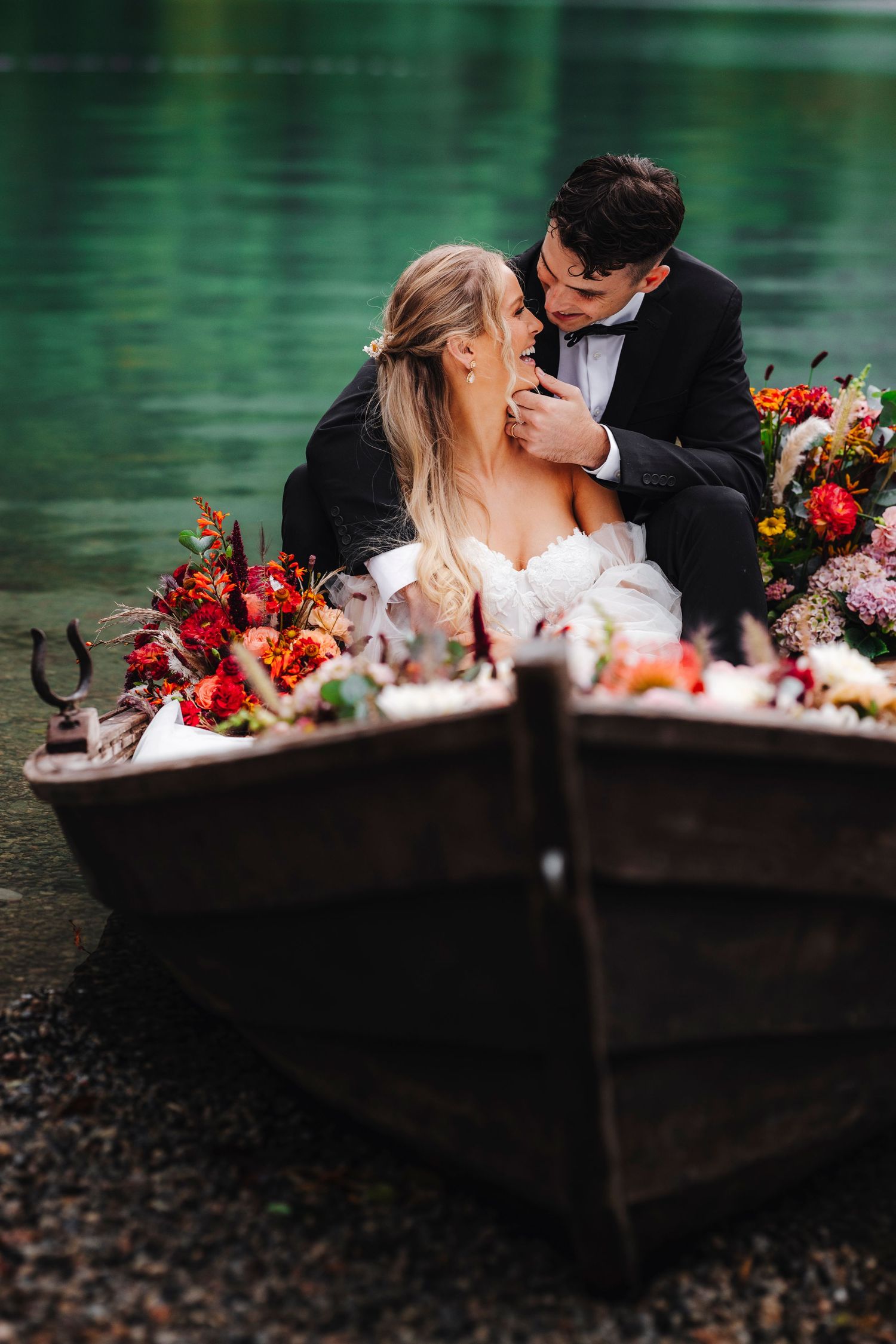 A newlywed couple shares a joyful moment near colorful floral arrangements by a waterfront.