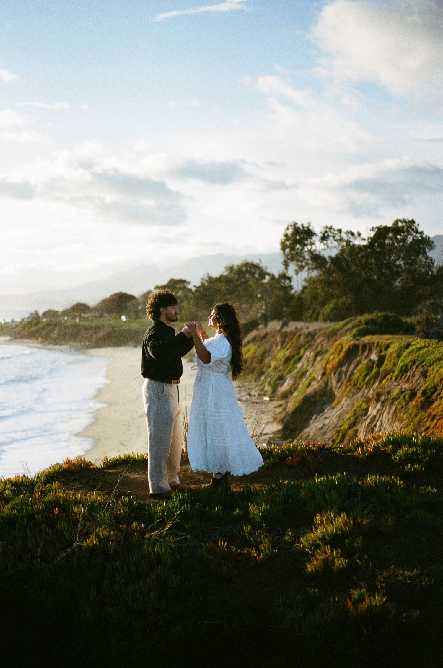 Two people stand on a coastal cliff overlooking the ocean at sunset with houses visible in the distance.