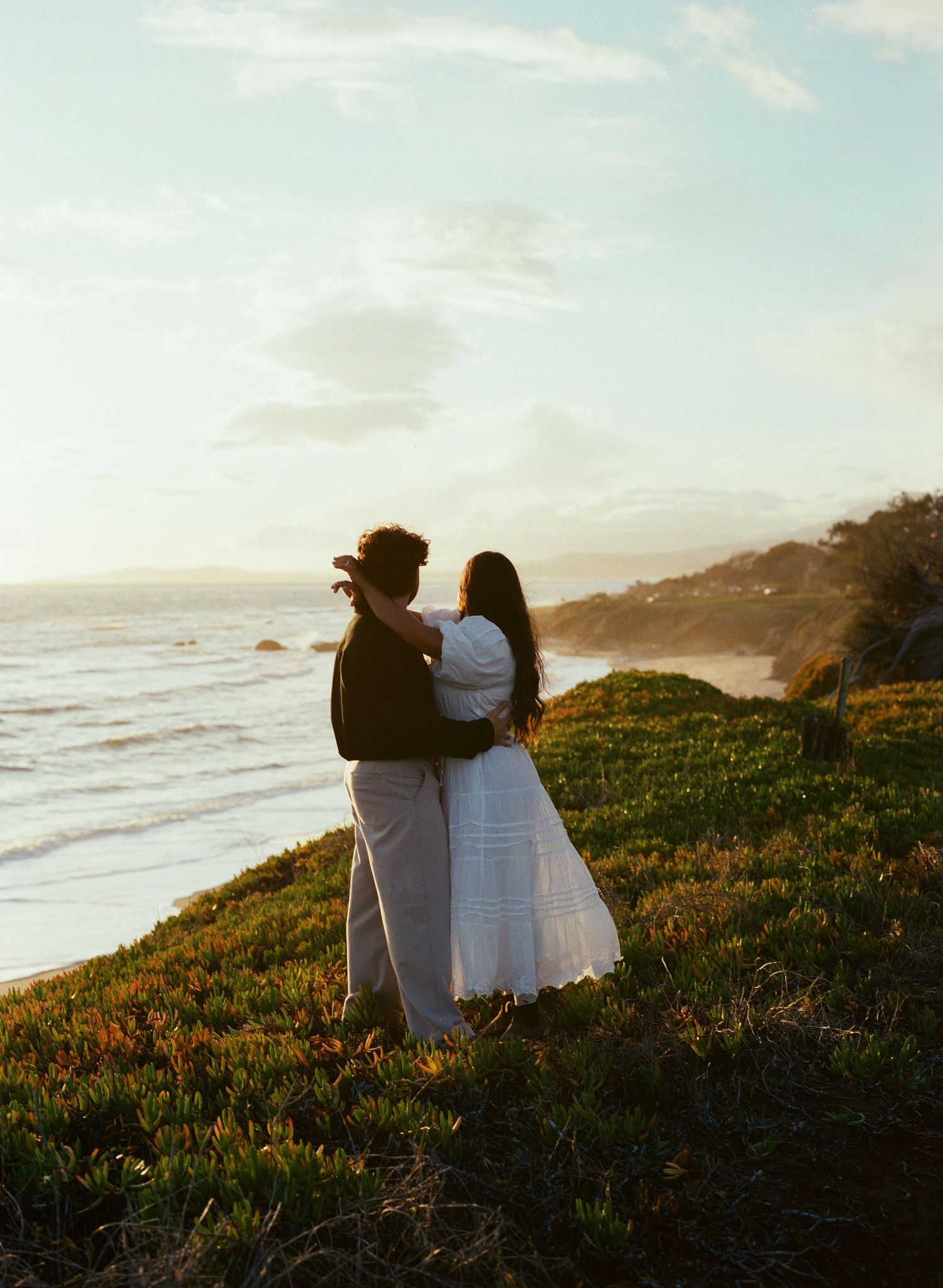 A couple stands together on a coastal cliff at sunset, overlooking the ocean in casual white and dark attire.