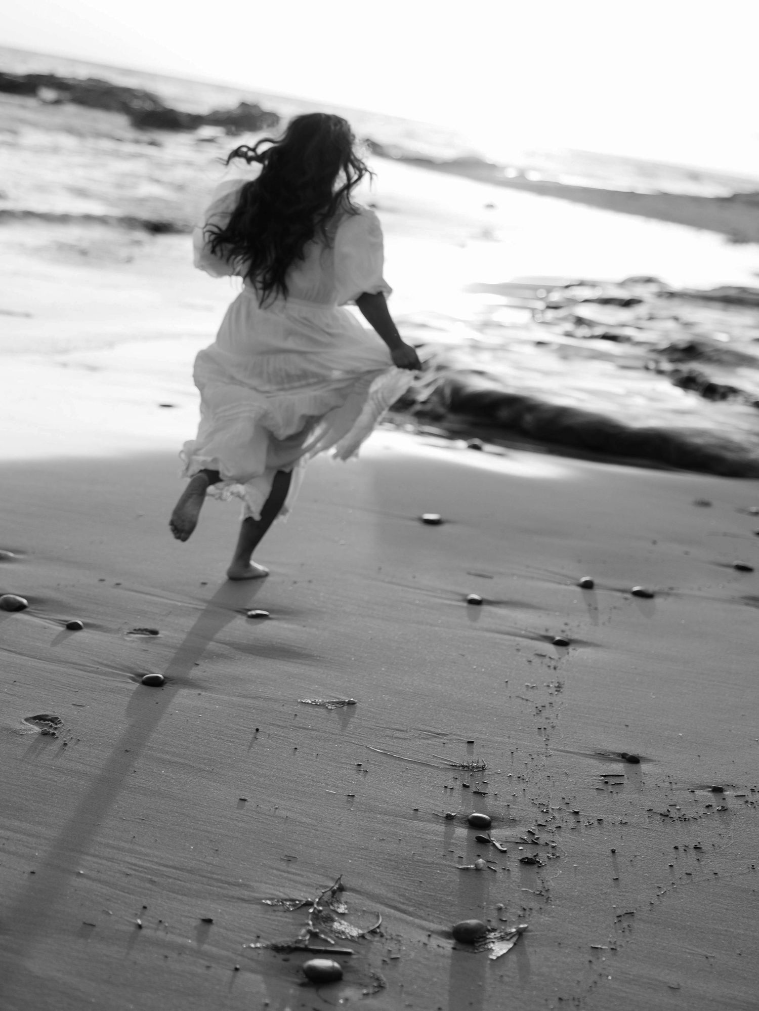 A person in a flowing white dress runs joyfully along a misty beach shore in black and white.