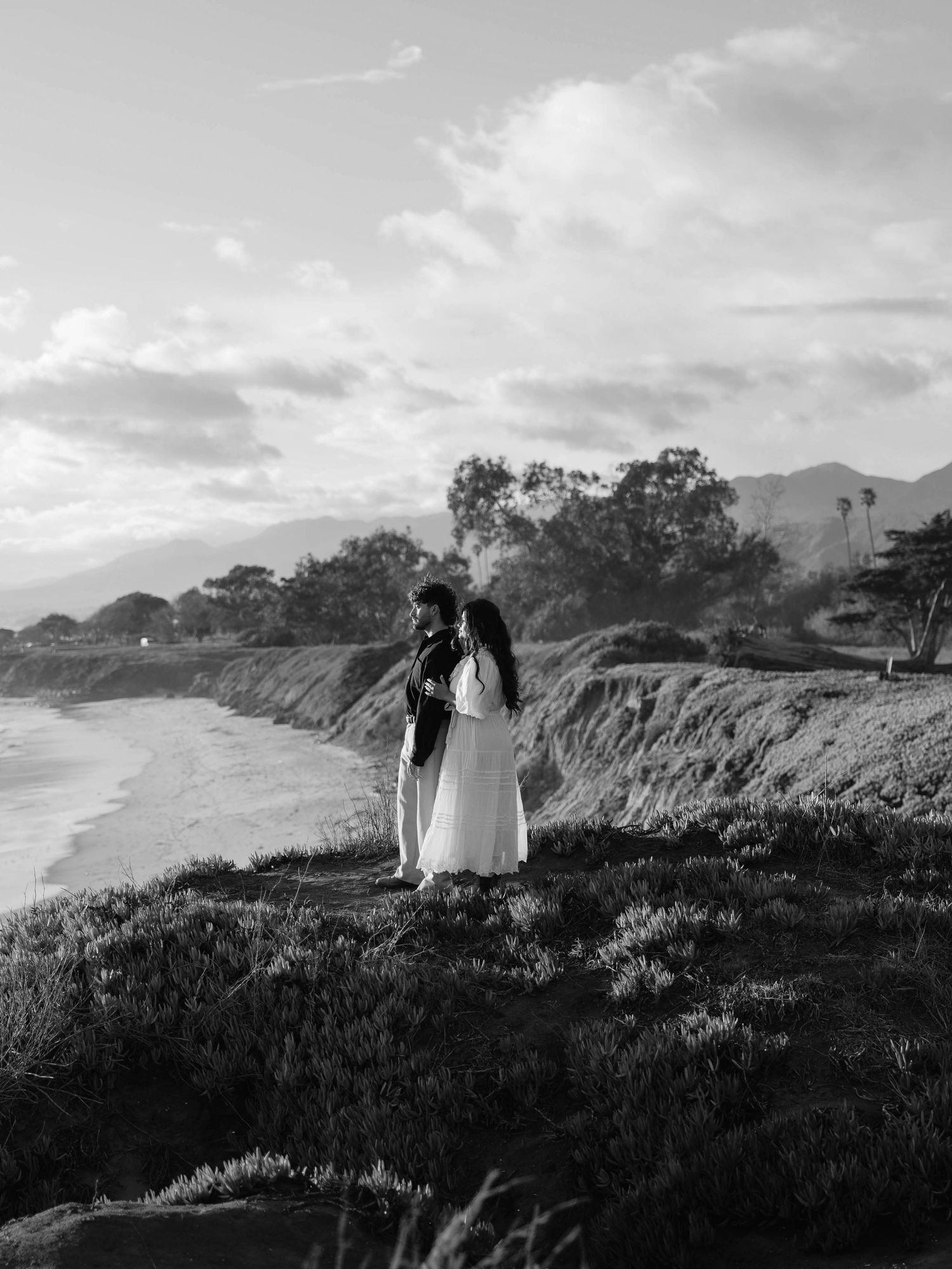 A black and white coastal landscape photo of a couple standing on a cliff overlooking the ocean at sunset.