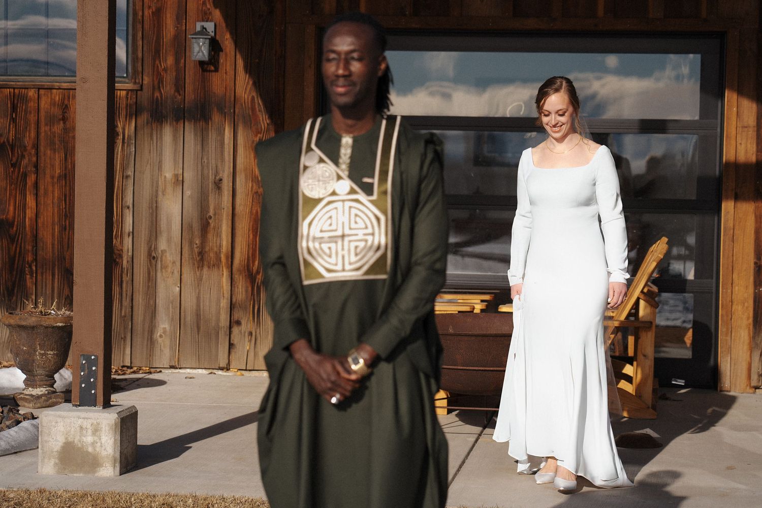 Two people in formal attire stand outside a rustic wooden building on a sunny day.