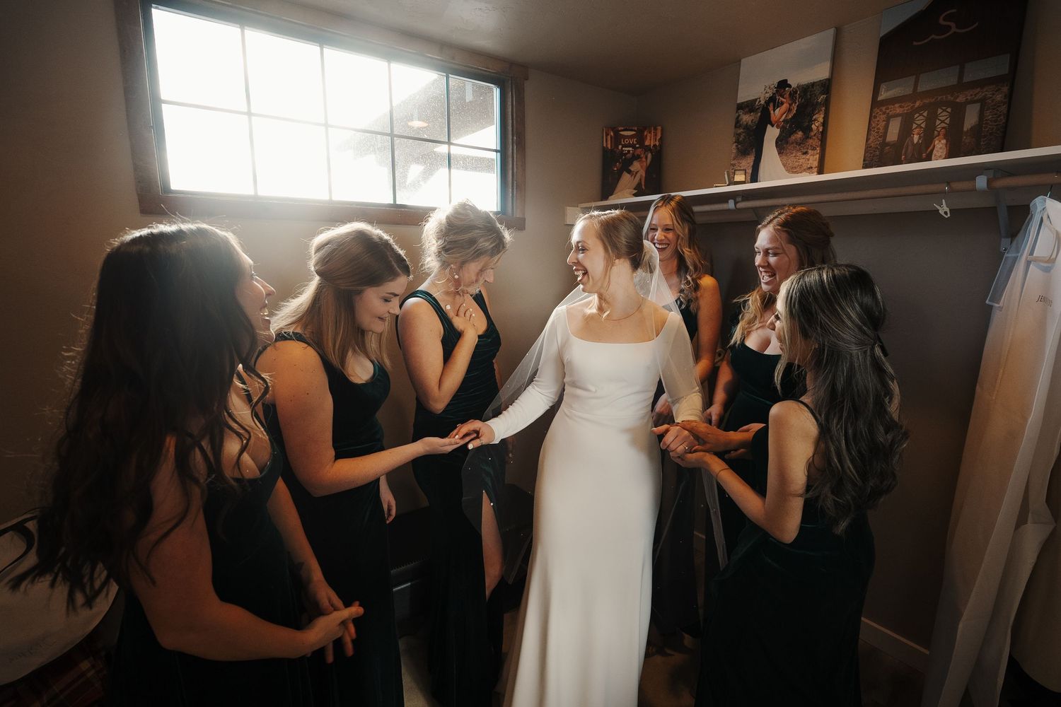 A bride in a white wedding dress shares a moment with her bridesmaids wearing black dresses in a sunlit room.