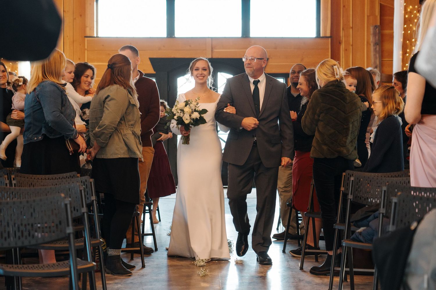 People gather in a rustic wooden venue for a wedding ceremony as the bride walks down the aisle.