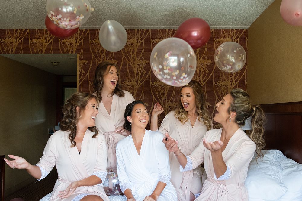 A bride and bridesmaids in white robes laugh while surrounded by pink and silver balloons at a pre-wedding gathering.