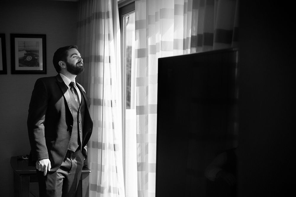 A suited man in black and white stands contemplatively by a window with soft light filtering through sheer curtains.