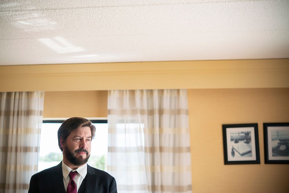 A man in a dark suit and red tie stands near curtained windows in a warmly lit room.