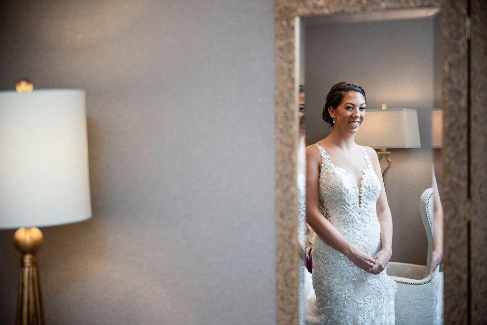 A bride in an elegant lace wedding dress reflected in a mirror in a softly lit room with table lamps creating a romantic atmosphere.