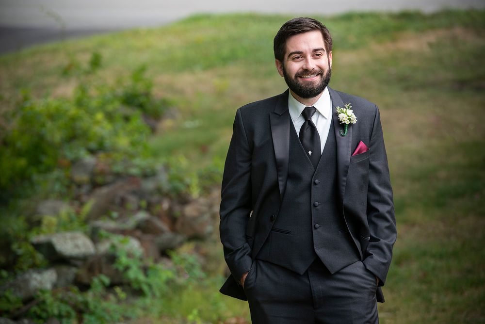 A groom wearing a dark three-piece suit with a burgundy pocket square poses on a green hillside.