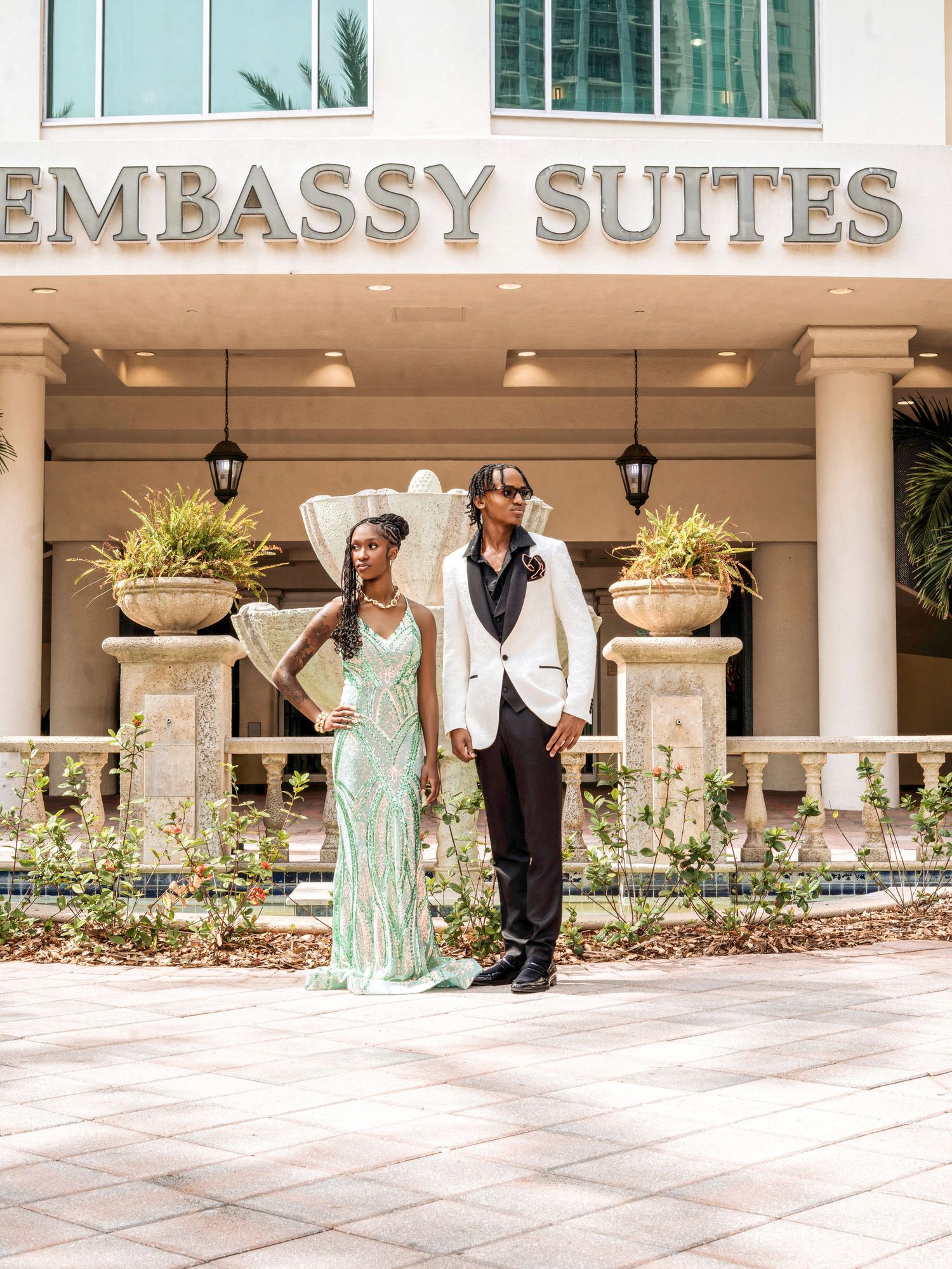 A couple poses outside Embassy Suites wearing formal attire with a mint green gown and white tuxedo jacket.