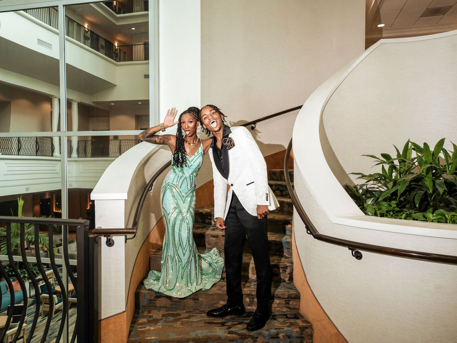 A festive couple in formal attire pose together on an elegant curved staircase in a hotel atrium.