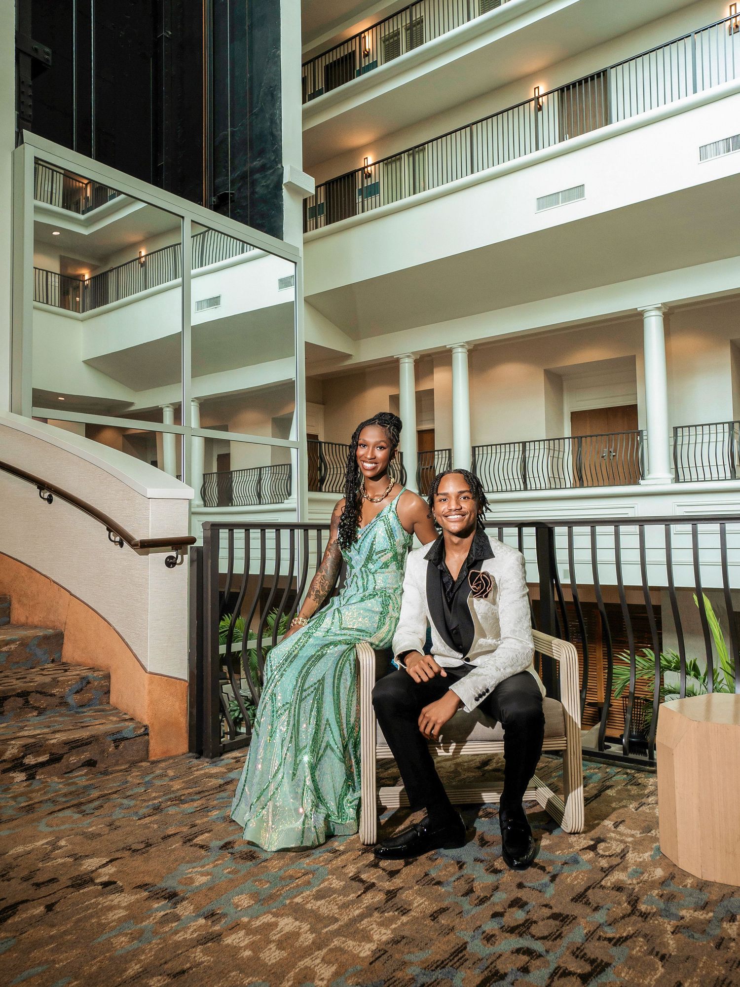 A couple in elegant formal attire poses on brick stairs in a hotel atrium with white balconies and columns.