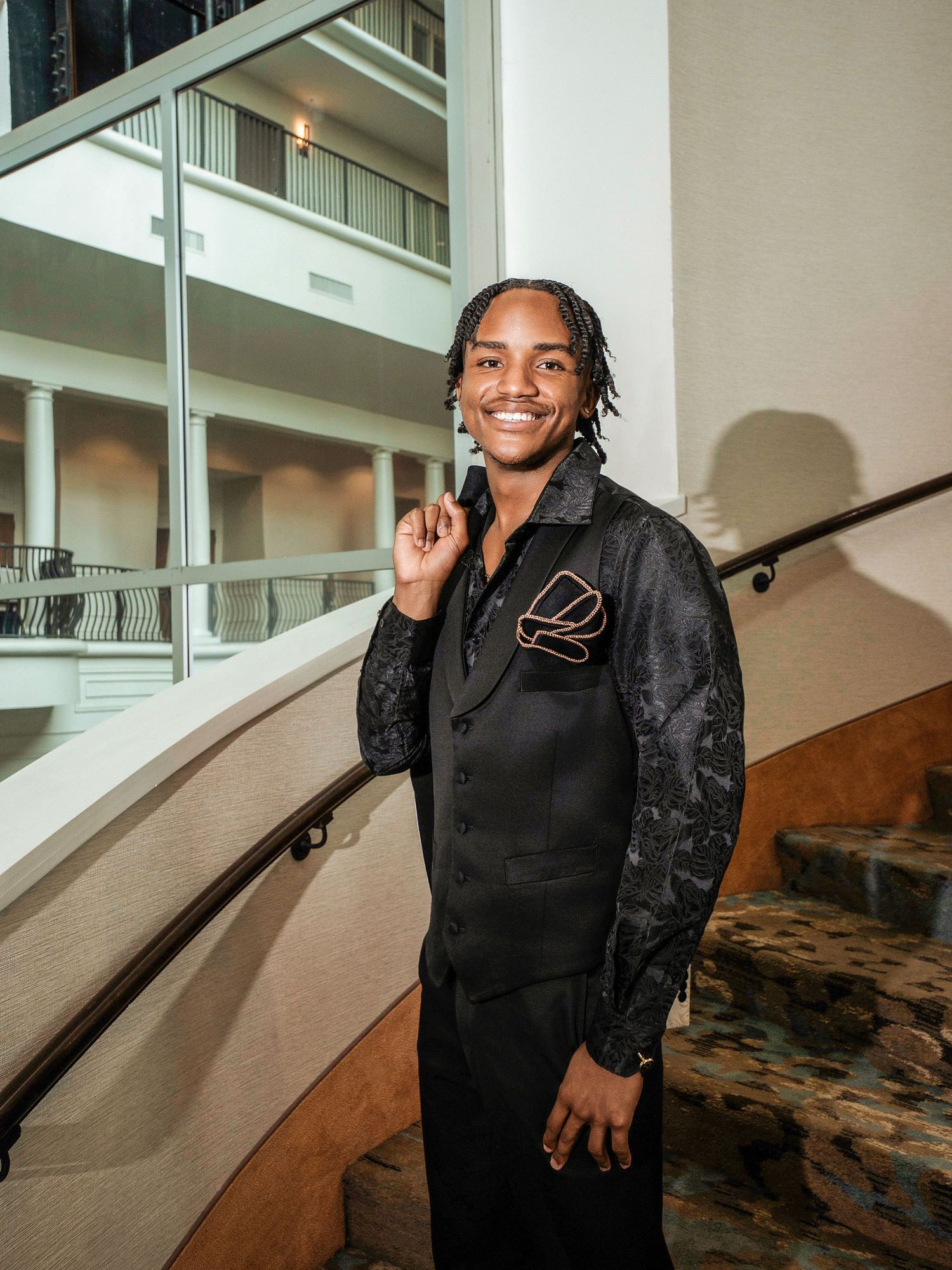 A well-dressed individual in a black suit with patterned sleeves stands on a curved staircase in an elegant interior.