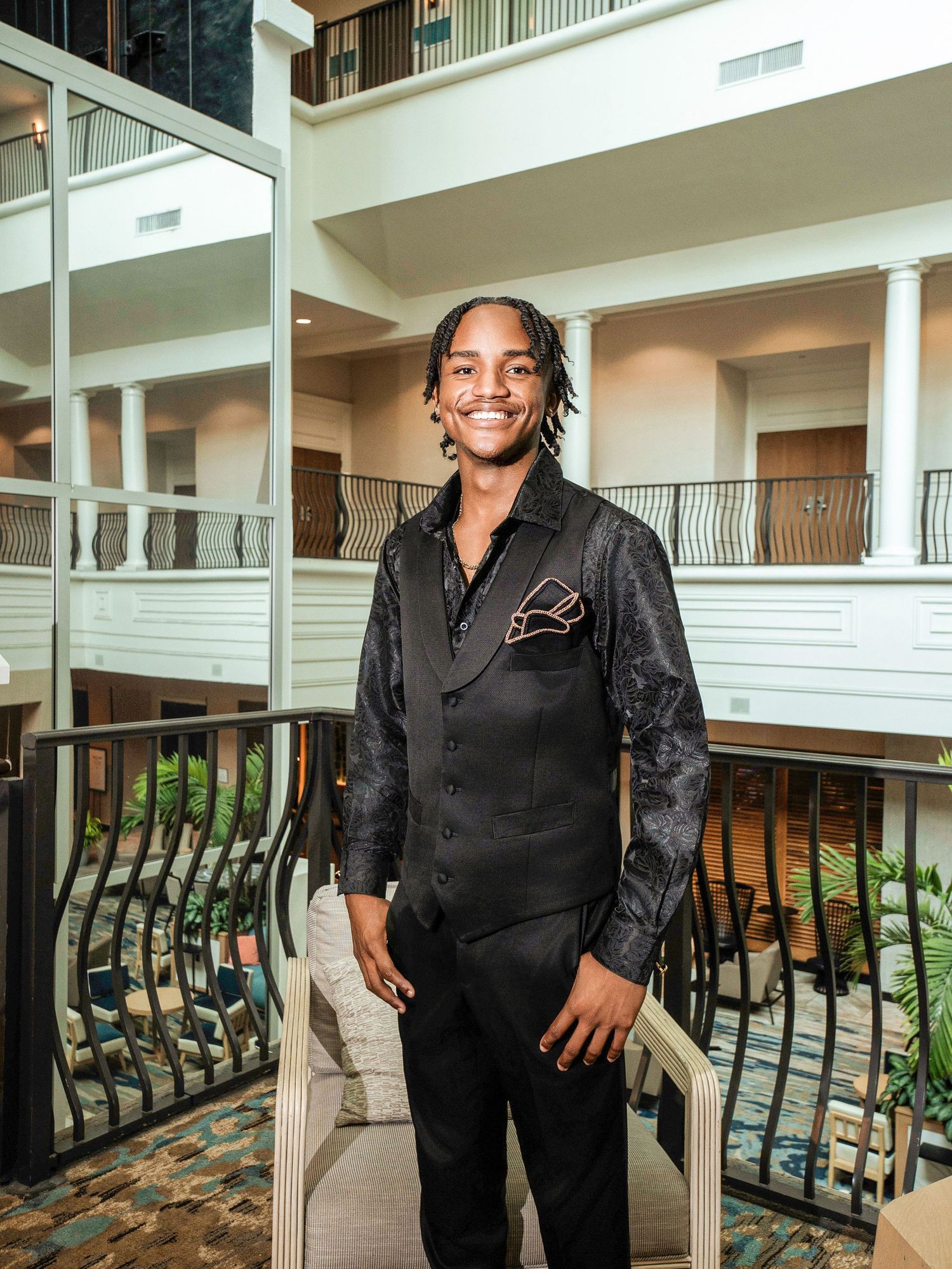 A person wearing a stylish black formal outfit stands by an interior balcony with white columns.