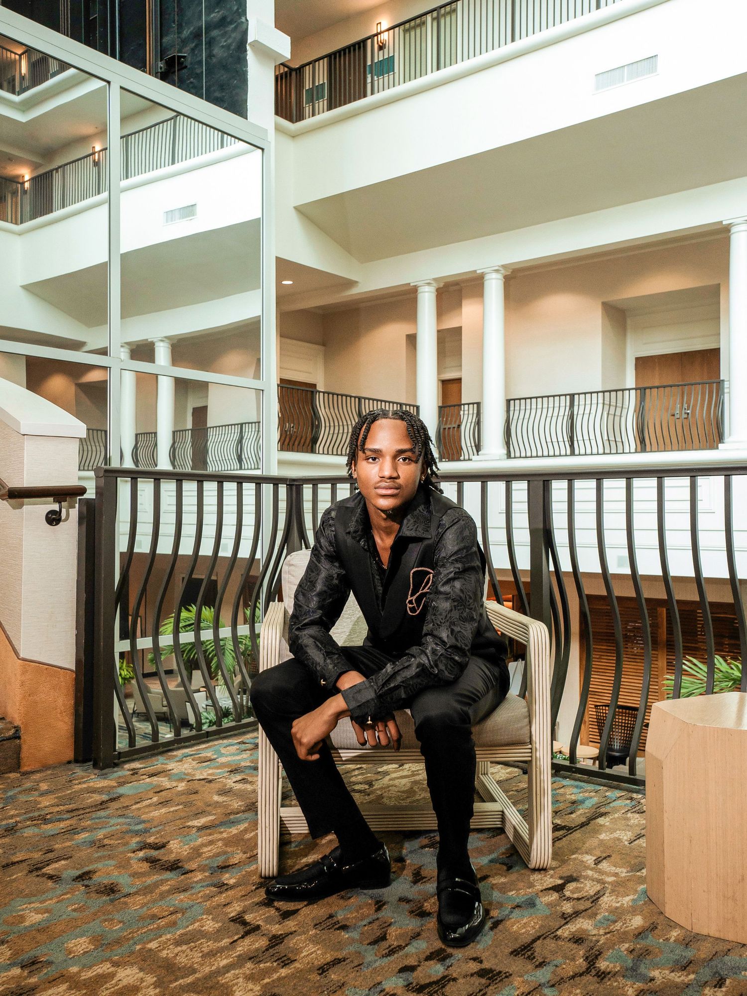 A stylishly dressed person sits on a chair inside an elegant interior space with balconies and columns.
