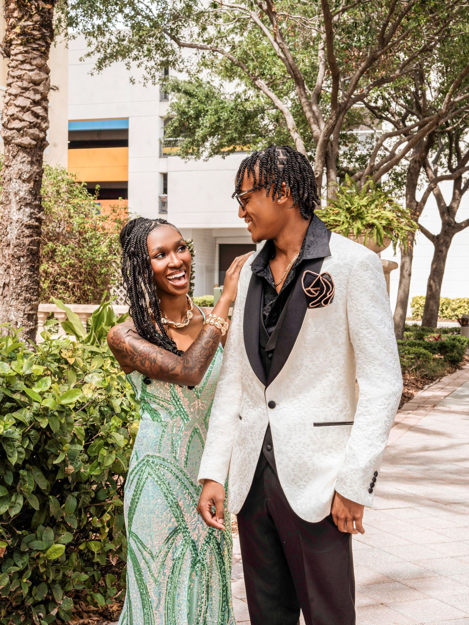A couple dressed in formal attire, with a white sequined jacket and mint green gown, share a happy moment outdoors.