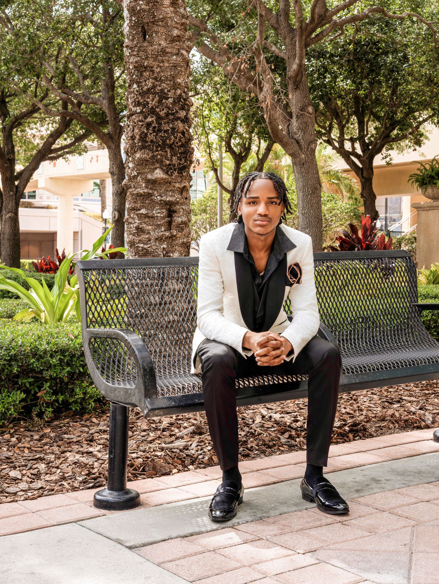 A person wearing a white blazer and black pants sits on a metal bench in a landscaped outdoor setting.