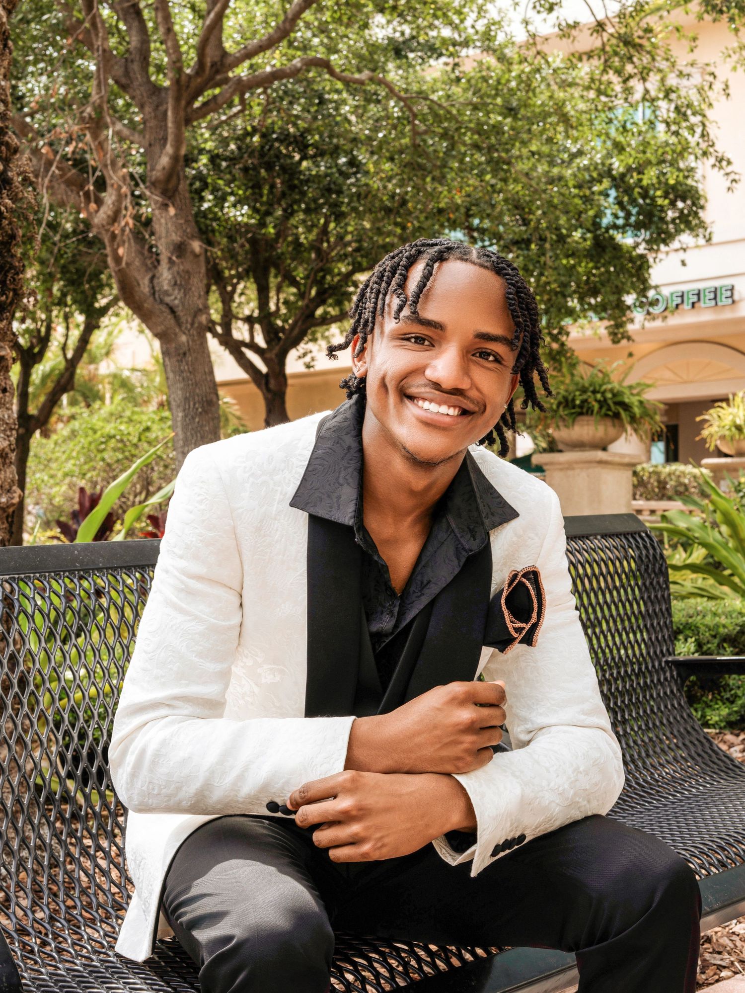 Young professional sitting on bench in white blazer and black shirt smiling in outdoor garden setting.