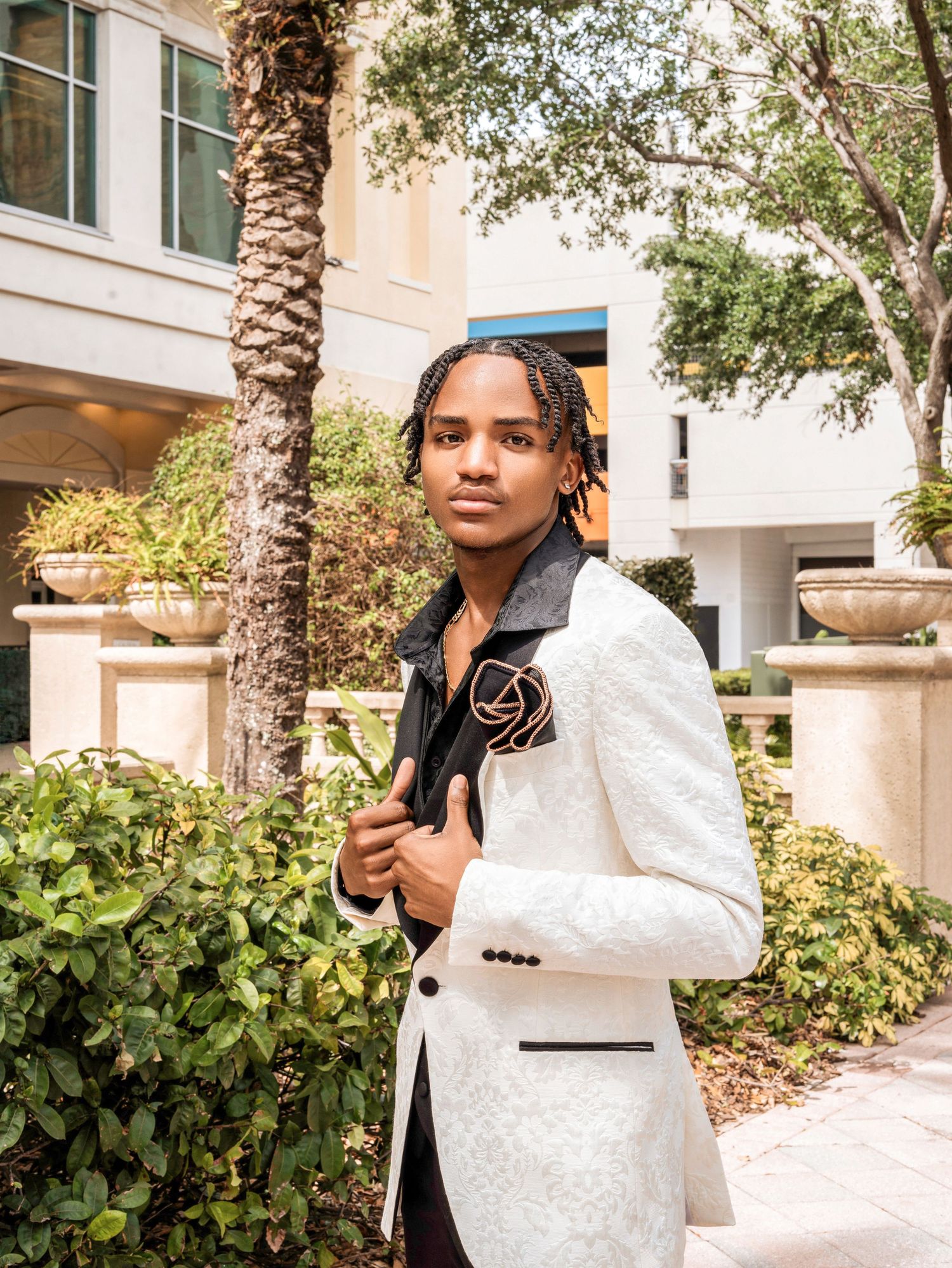 A person in an elegant white dinner jacket poses outdoors against a backdrop of tropical foliage and architecture.