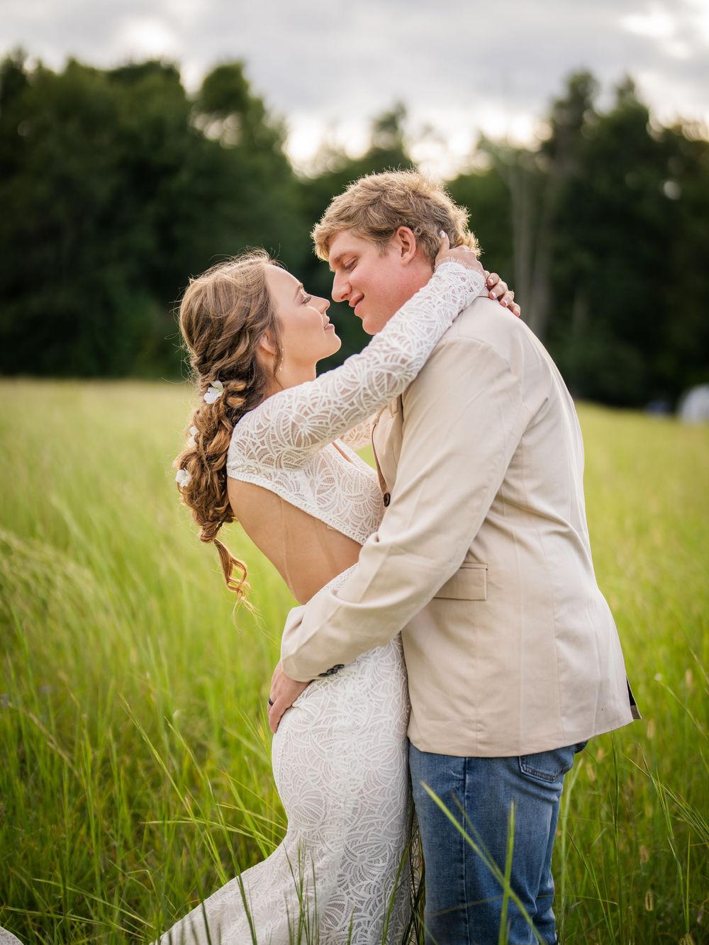 Two people share a romantic embrace in a tall grass field with trees in the background during their wedding photoshoot.