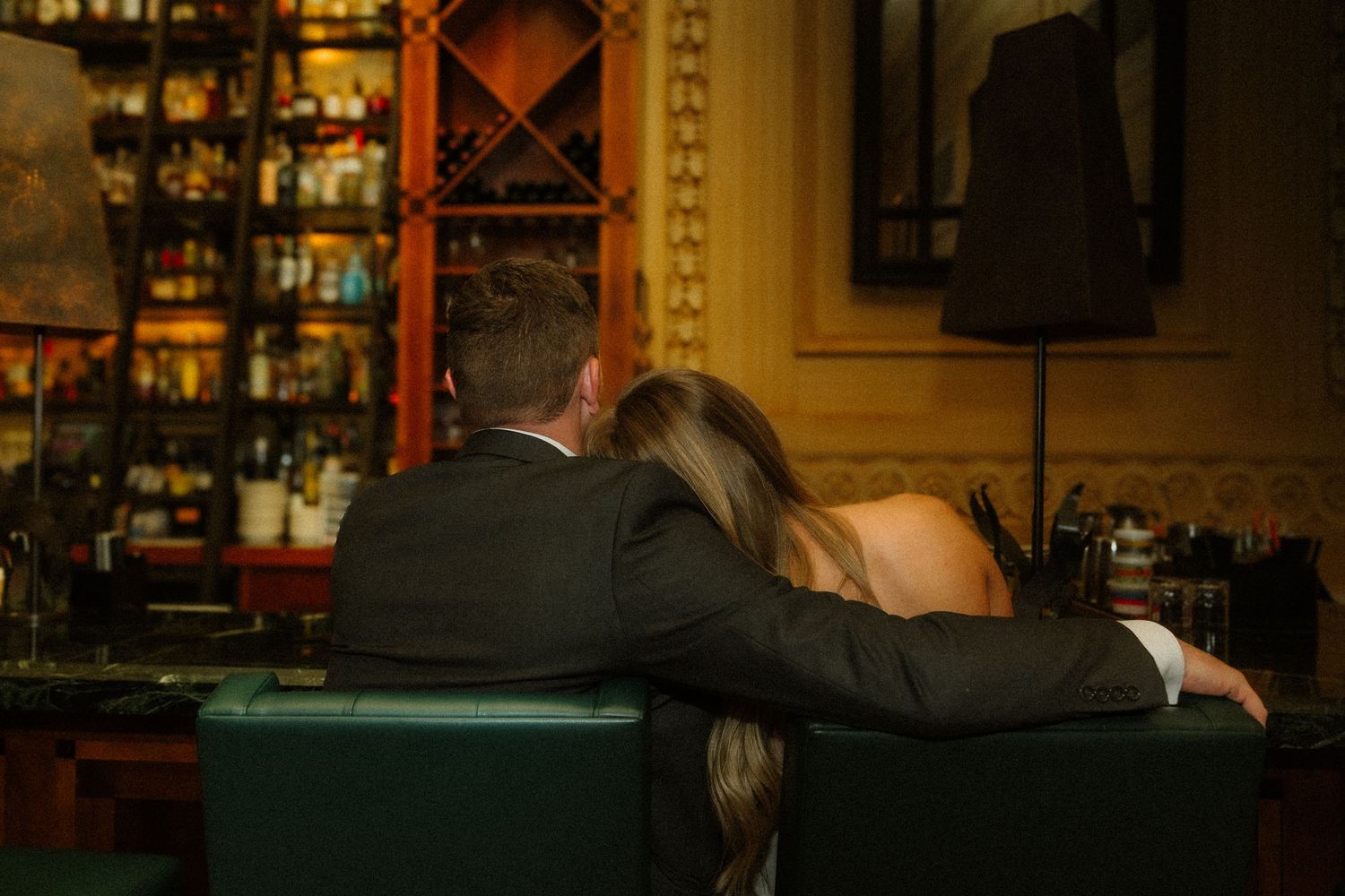 A couple sits intimately at a dimly lit bar with bottles and glasses visible in the background.
