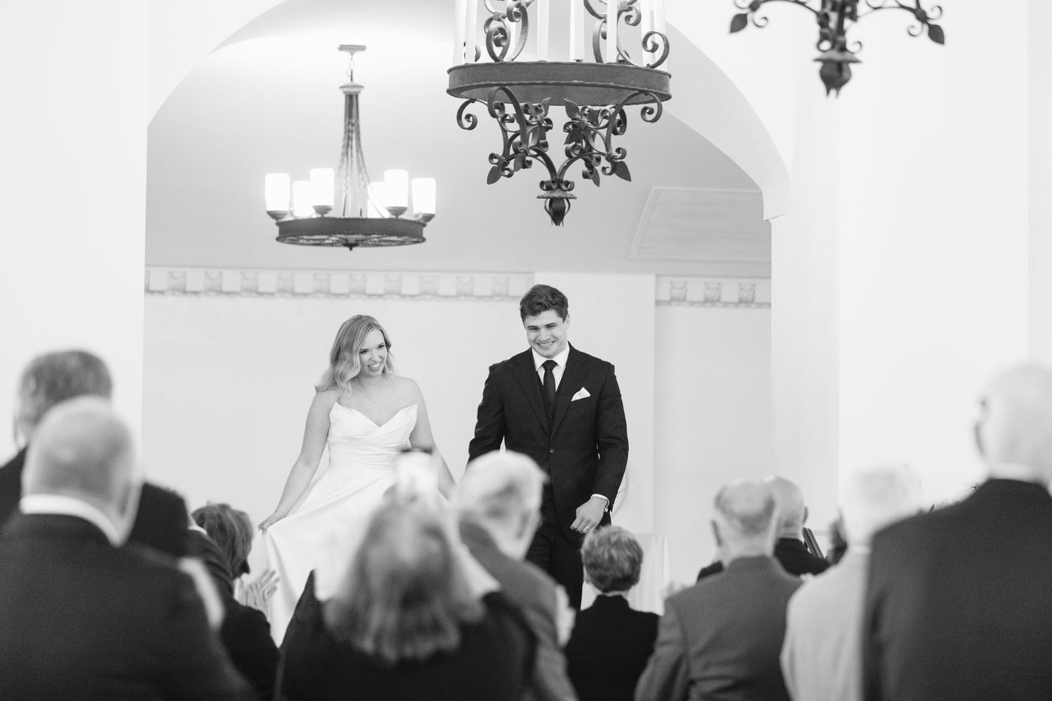 A black and white wedding ceremony photo showing a couple standing at the altar under an ornate arch with guests seated.
