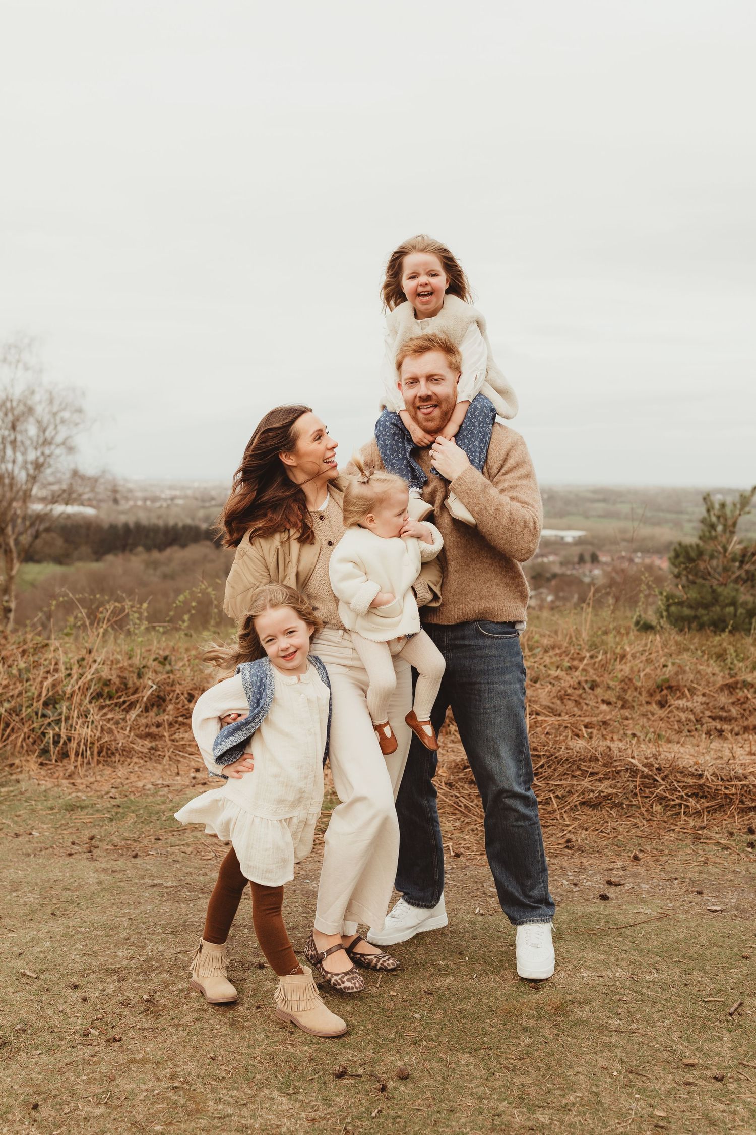 A joyful family wearing coordinated neutral outfits poses together outdoors on a hilltop during autumn.