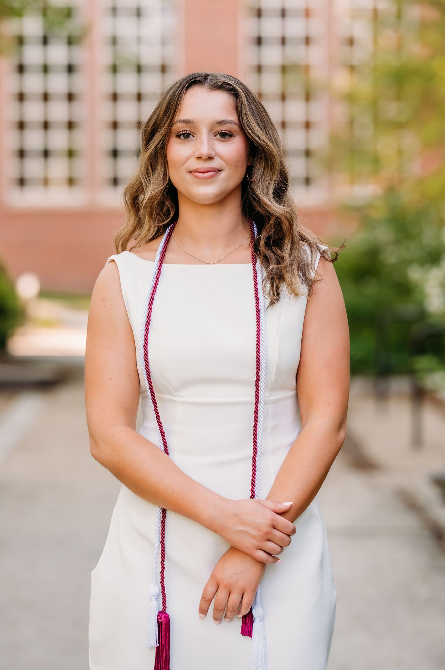 A graduate wearing a white sleeveless dress poses outdoors with a red graduation tassel hanging around their neck.